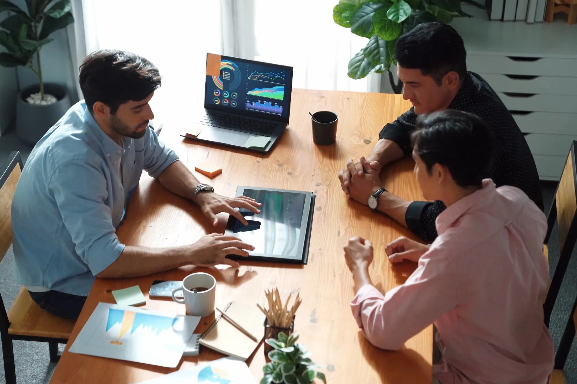 A man at a meeting table talking two colleagues through figures on a tablet while they lean in to follow