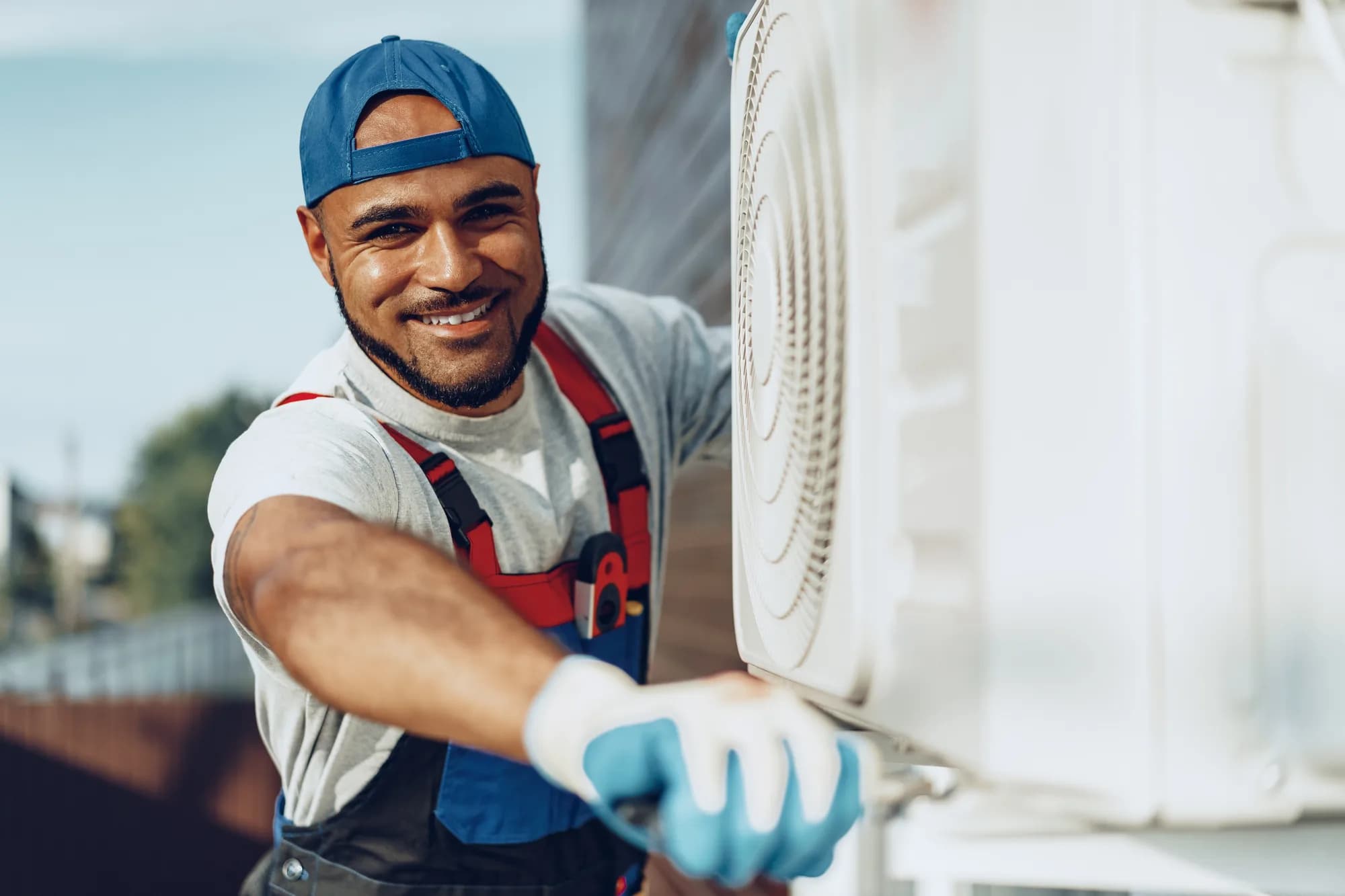 A smiling HVAC technician in a cap and work overalls servicing an outdoor air-conditioning unit