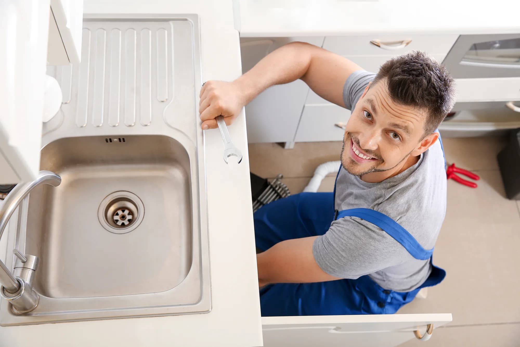 A smiling plumber in blue overalls seated beneath an open kitchen sink cabinet, holding a spanner and looking up