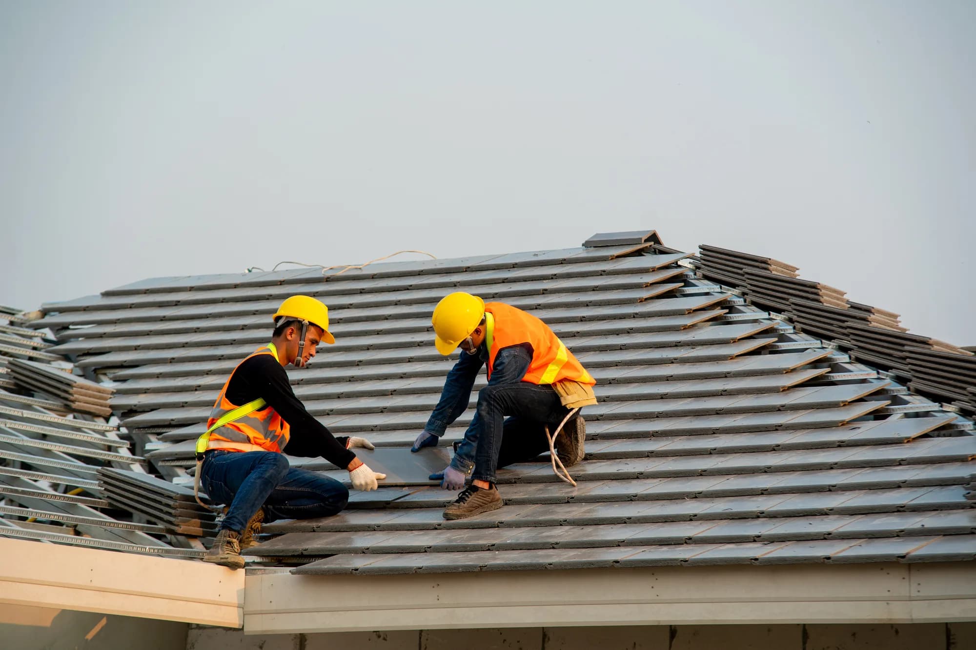Two roofers in hard hats and hi-vis vests kneeling on a pitched roof, laying gray concrete tiles
