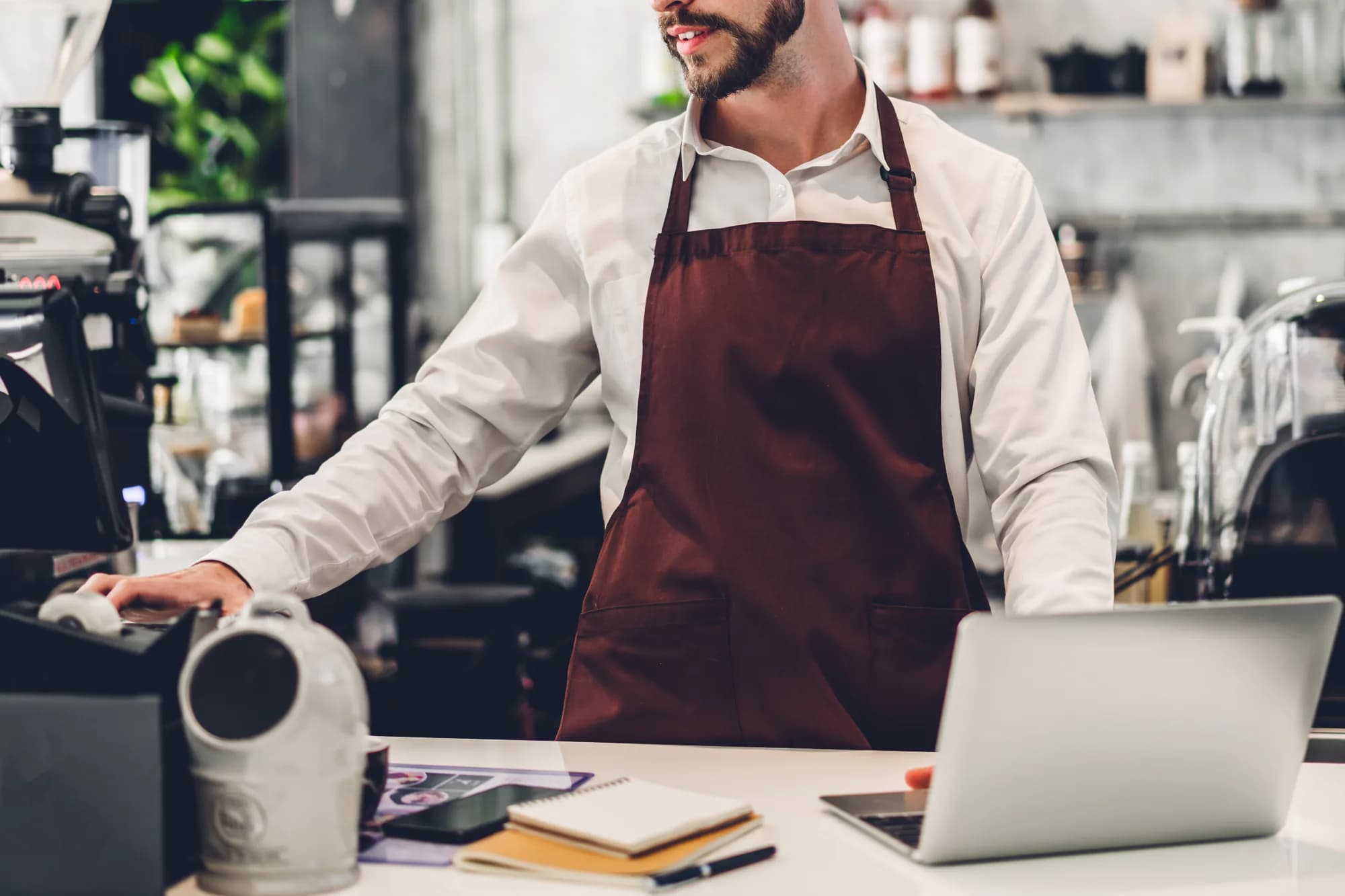 A small business owner in an apron at a cafe counter, one hand on a computer mouse by the till and the other at an open laptop, with a notebook and pen beside it